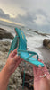 Model holding aqua embellished heel by the ocean